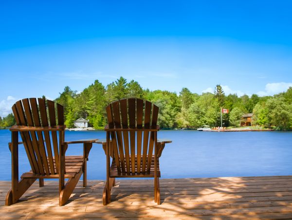 Two wooden chairs on a dock overlooking a calm lake with trees and a Canadian flag in the distance.