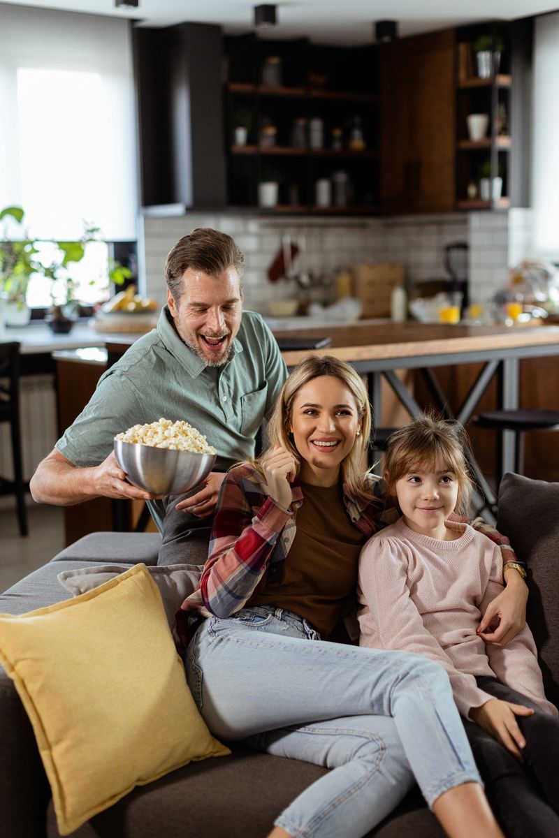 A family of three is comfortably nestled on a couch, their faces reflecting excitement and attentiveness as they share a bowl of popcorn during a suspenseful movie night