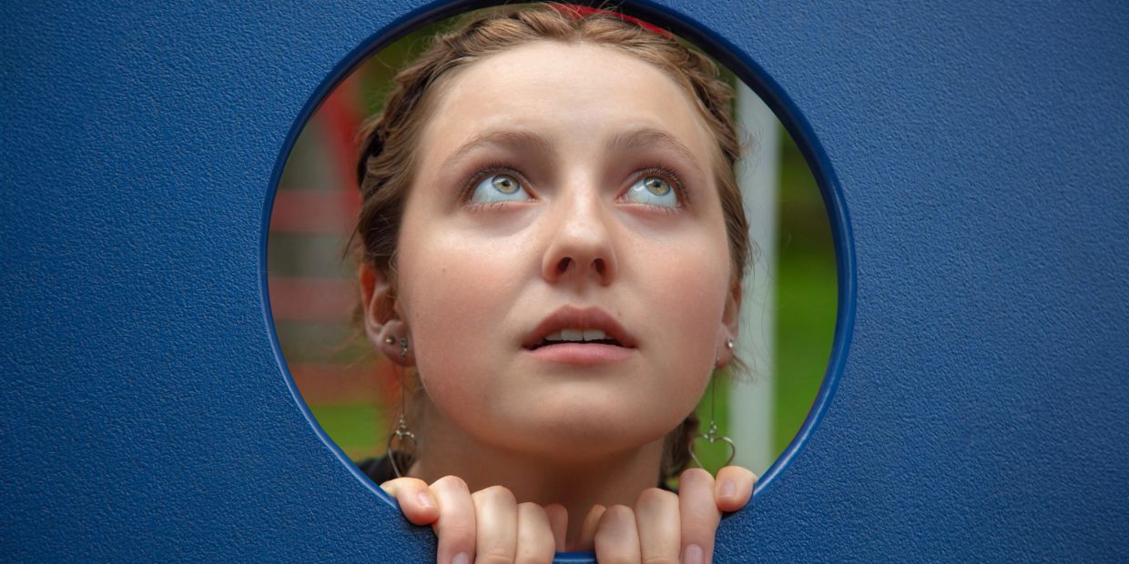 Young woman looks upwards through a circular blue frame, hands gripping the edge.