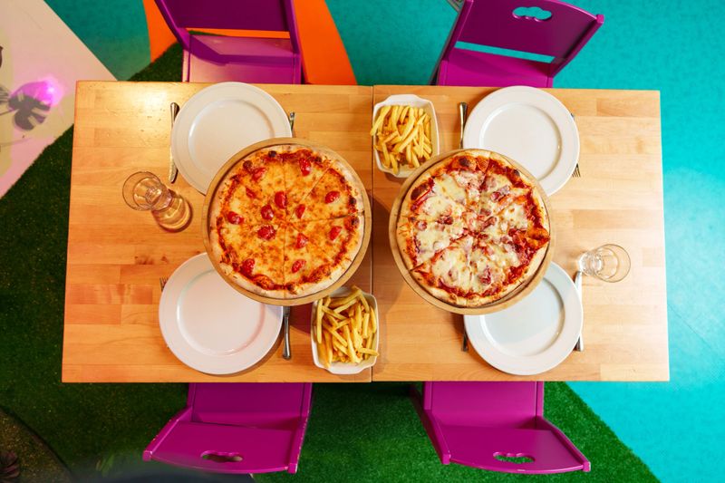 Two delicious pizzas with golden-brown cheese are displayed on elevated wooden stands above a table adorned with a colorful tablecloth. Beside the pizzas, there is a bowl of golden French fries and glasses filled with amber-colored beverages, suggesting an informal outdoor meal.