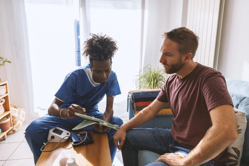 A healthcare worker in blue scrubs sitting on an orange stool, attentively using a digital tablet during a medical consultation with a patient in a bright, welcoming living room.