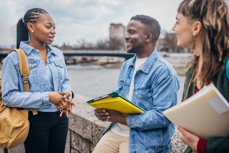 Diverse group of college students from different cultural backgrounds enjoying a casual conversation and study group outdoors by the scenic river on campus. Dressed in casual attire