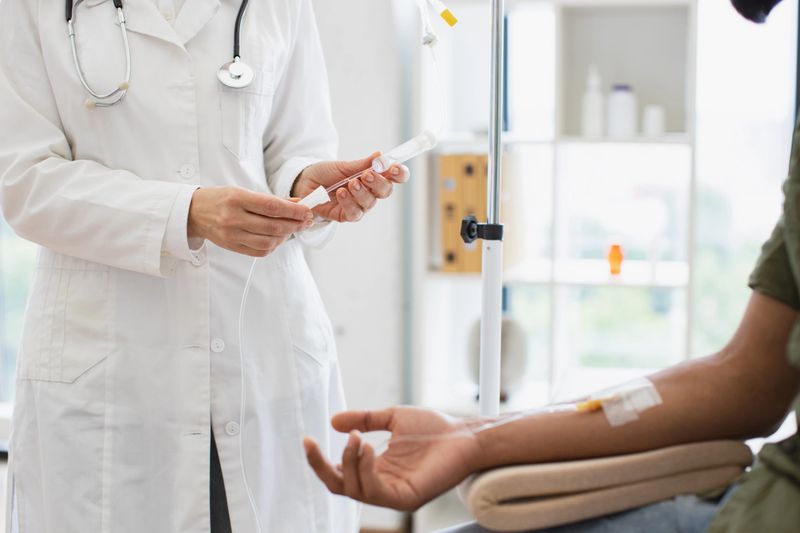 Young man in casual wear taking dropper while mature female making system review. Efficient general practitioner checking solution for chemotherapy of male sitting on exam couch.