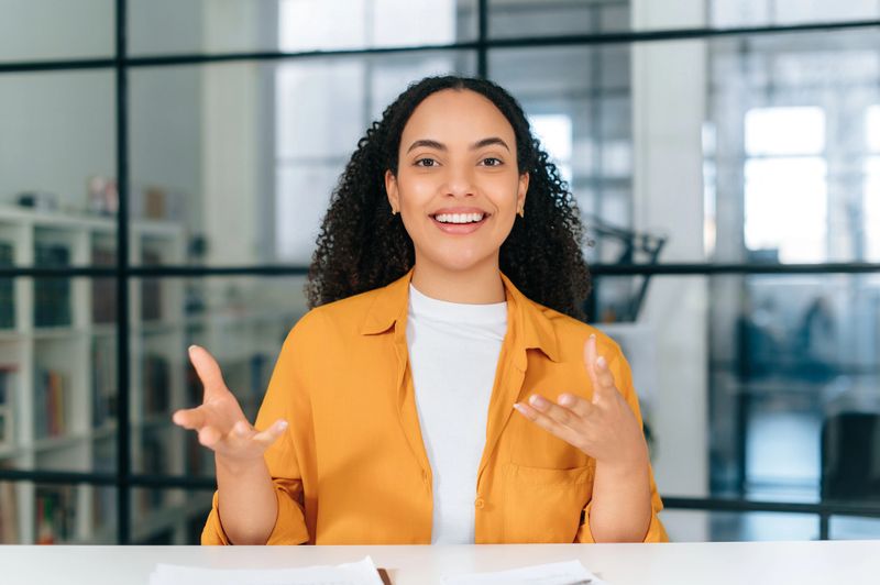 Confident pretty hispanic or brazilian curly haired young woman, talking via video call, listens or conducts an online lesson, coaching session, lecture, brainstorm, gesturing with hands, smiles