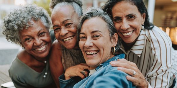 Four women smiling and taking a close-up selfie together outdoors.