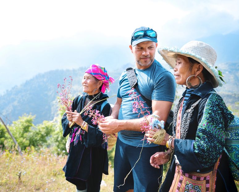 Three people holding flowers outdoors in a mountainous area.