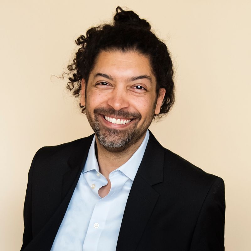 Portrait of stylish modern professional multiracial man on beige background. He is in his forties, wearing a light blue shirt and a black suit. He has long curly hair tied in a man bun and a beard. He is standing and looking at the camera. Vertical head and shoulder studio shot with copy space. This was taken in Montreal, Quebec, Canada.
