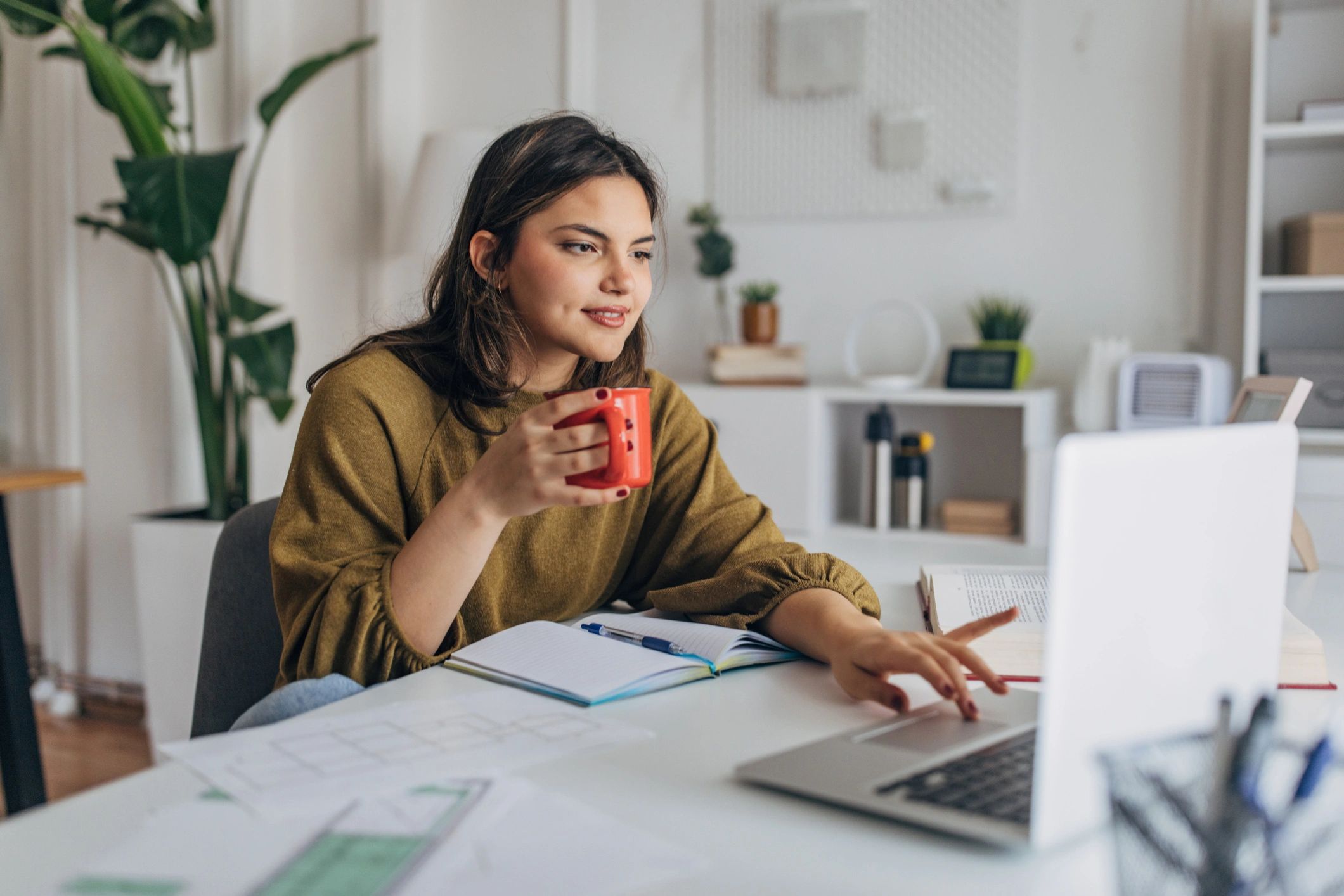 woman working on a laptop with a notebook in front of her, drinking a cup of coffee
