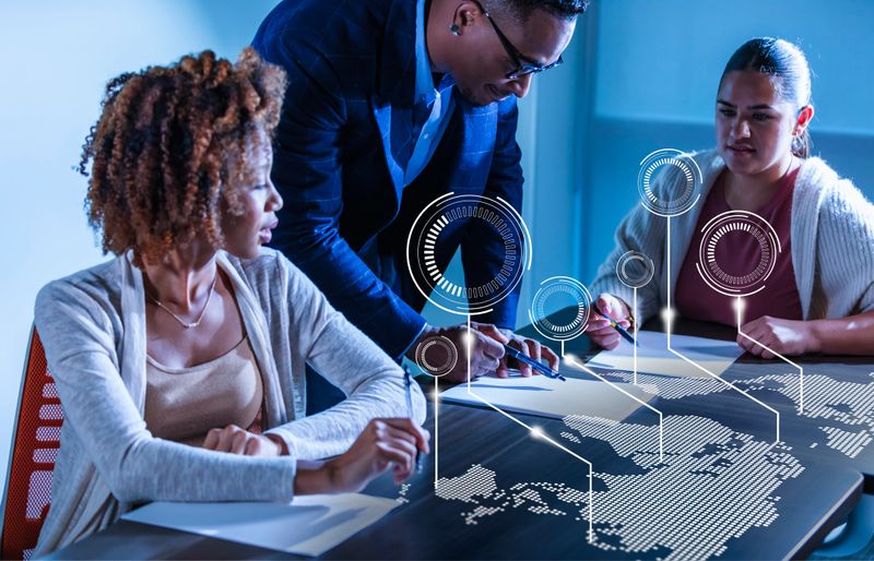 A multiracial group of three business people in a meeting to discuss global business strategy. An African-American man is standing between two young women seated at the conference table looking at papers. A graphical representation of a world map is overlayed on the table in front of them.