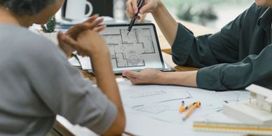 Two people discussing a house floor plan on a tablet and paper at a table.