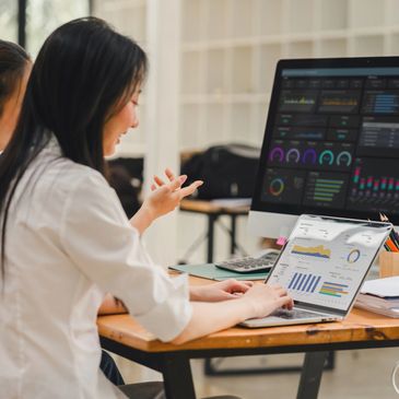 Two women analyzing colorful data charts on laptop and desktop screens in a modern office.