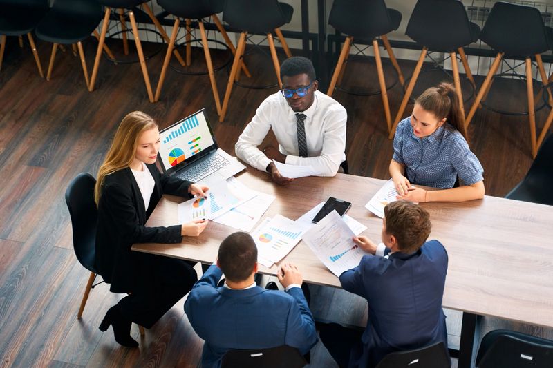 Multiracial team discusses strategy around table with papers, graphs. Female CEO leads business meeting, analyzes charts on laptop in modern office. Focus on collaboration, corporate growth.
