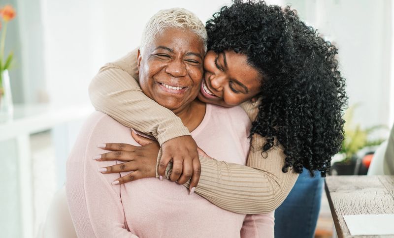 African daughter hugging her mum indoors at home - Main focus on mother face - Mom day and family love concept
