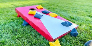 A red and white cornhole board with yellow and blue bean bags on grass.