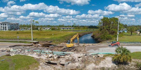 Construction machinery repairing a damaged road over a water canal under a blue sky.