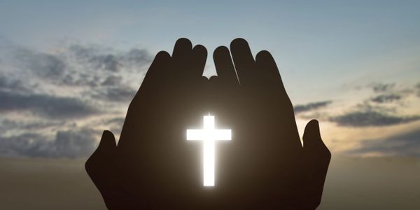 Hands holding a glowing cross against a sunset sky.