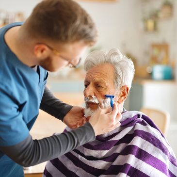 A caregiver shaves an elderly man's face with care.