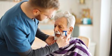 A caregiver shaves an elderly man's face with care.