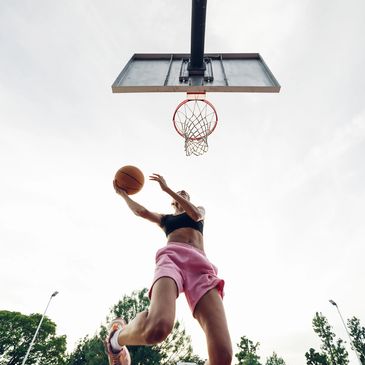 Person in pink shorts jumps to shoot a basketball at an outdoor court.
