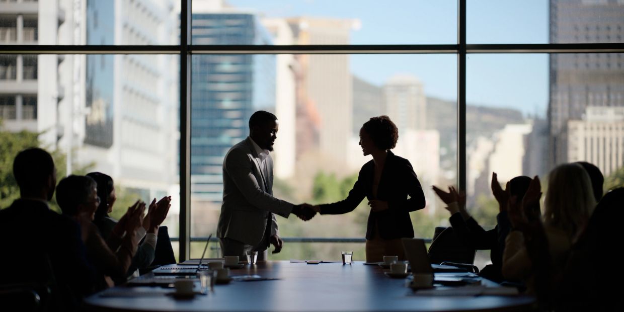 Two professionals shaking hands in a meeting room with applauding colleagues.