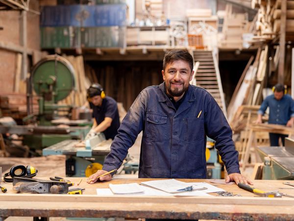 Smiling carpenter standing at a workbench in a busy woodworking shop.