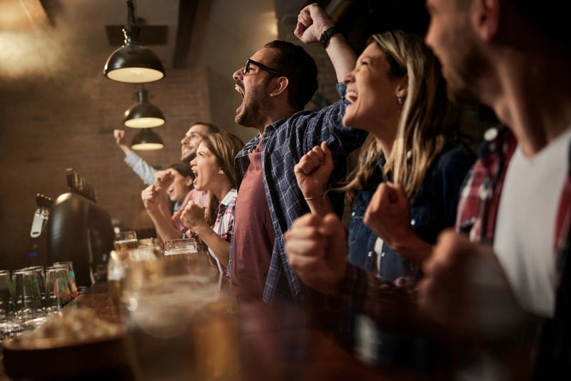 Cheerful fans having fun while celebrating the success of their sports team while watching a game on TV in a bar. Focus is on man with eyeglasses.