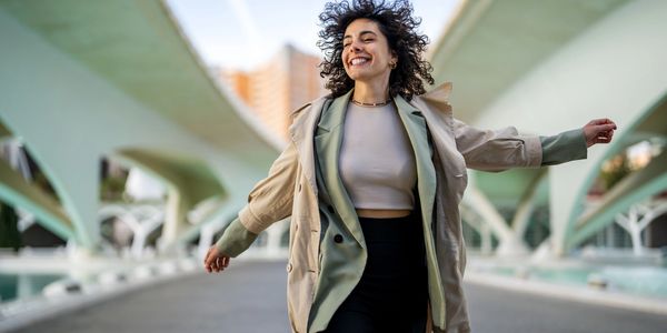 Joyful woman with curly hair walking under a modern bridge, smiling brightly.