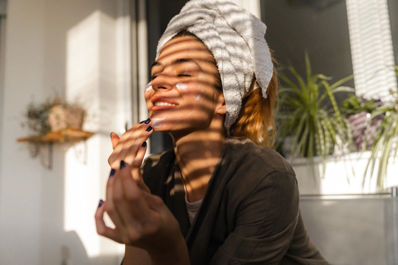 Young beautiful woman is sitting on the balcony nourishing her face with a hydrating cream. Woman has just washed her hair and has a towel on her head. Shadow striper are falling on beautiful woman. Woman selfcare at home.