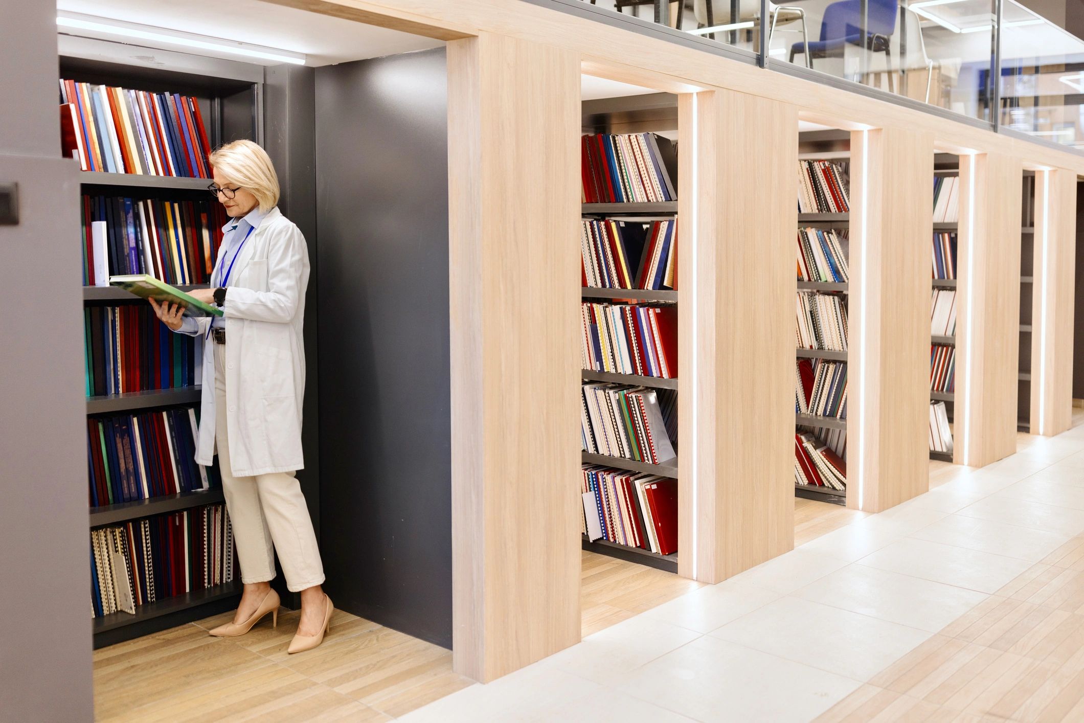 Woman in white coat reading a book in a modern library.