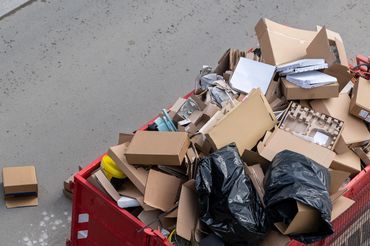 A red dumpster filled with cardboard boxes and black garbage bags on a street.