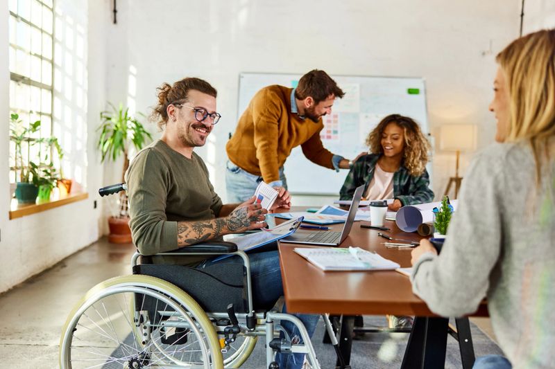 Happy creative man in wheelchair communicating with his colleague while having a meeting at casual office.