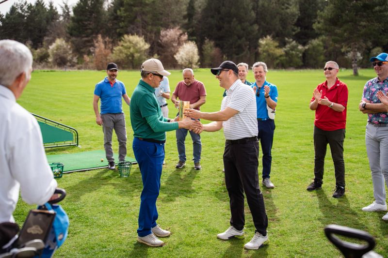 Golfer proudly receiving a trophy, after winning a golf tournament. Fellow golfers are standing in the background cheering him on.