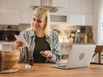 Woman reading medication instructions while sitting at a kitchen table with a laptop.