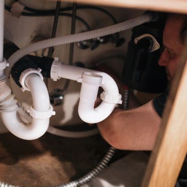 Gloved hand fixing a white plastic drain pipe under a sink.