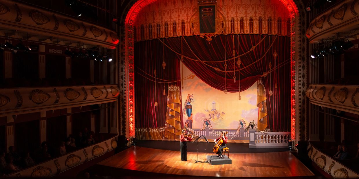 Two musicians perform on an ornate stage with red curtains.