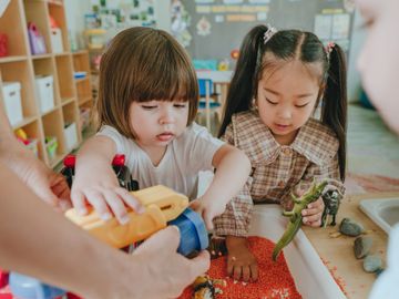 Two young children playing with toy animals and a toy truck in a sensory bin.