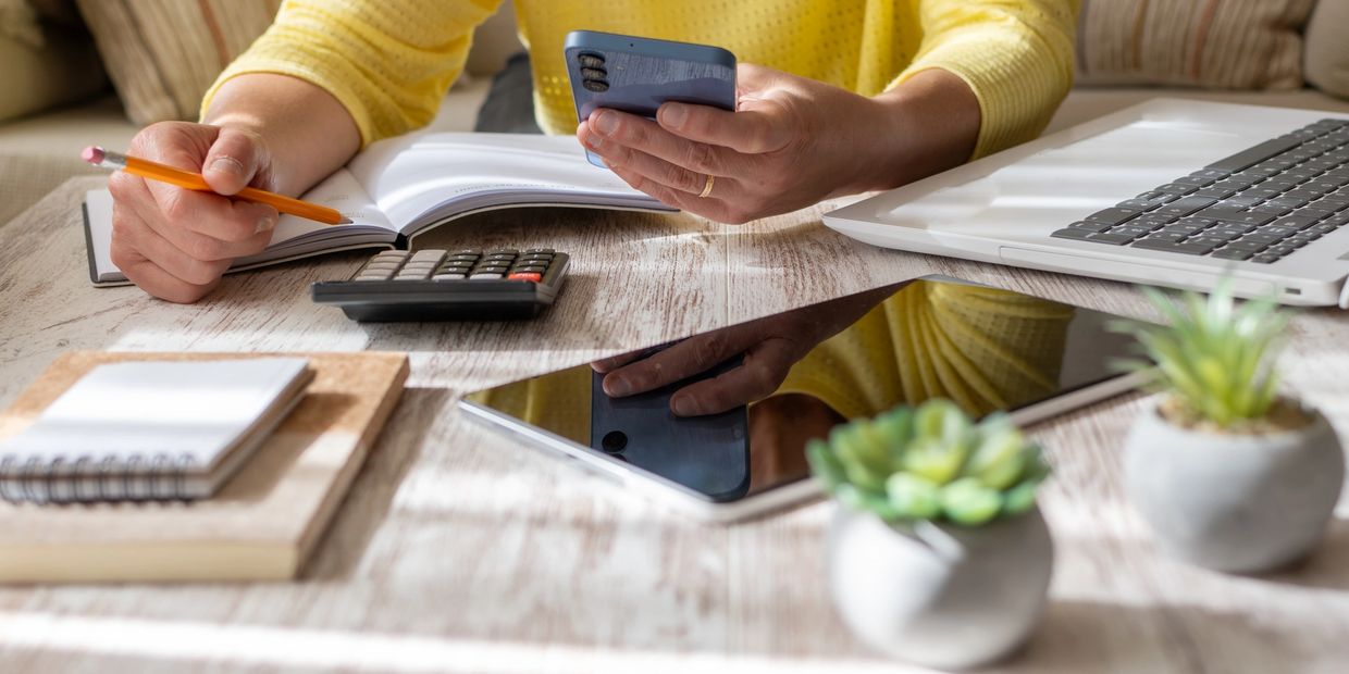 Person budgeting at home with calculator, phone, and laptop on a wooden desk