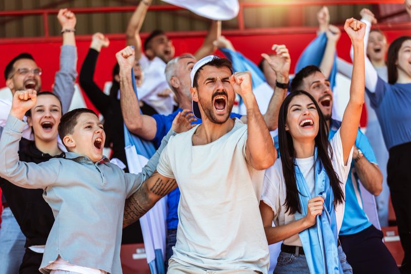 Sport fans cheering at the game on stadium. Wearing blue and white colors to support their team. Celebrating with flags and scarfs.