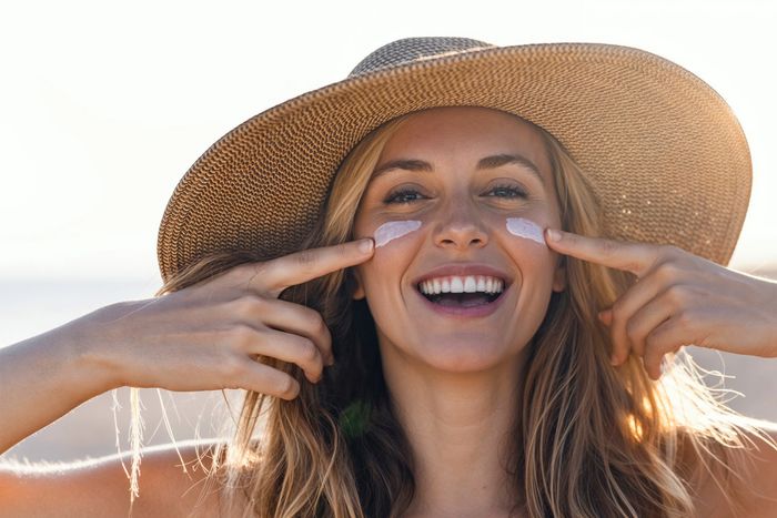 Smiling woman applying sunscreen on her cheeks at the beach wearing a sun hat.