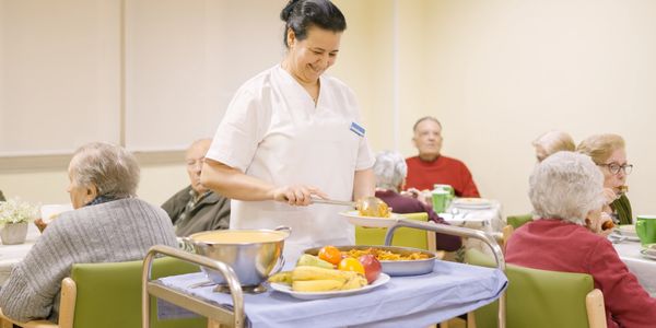 Caregiver serving food to elderly people in a nursing home dining room.