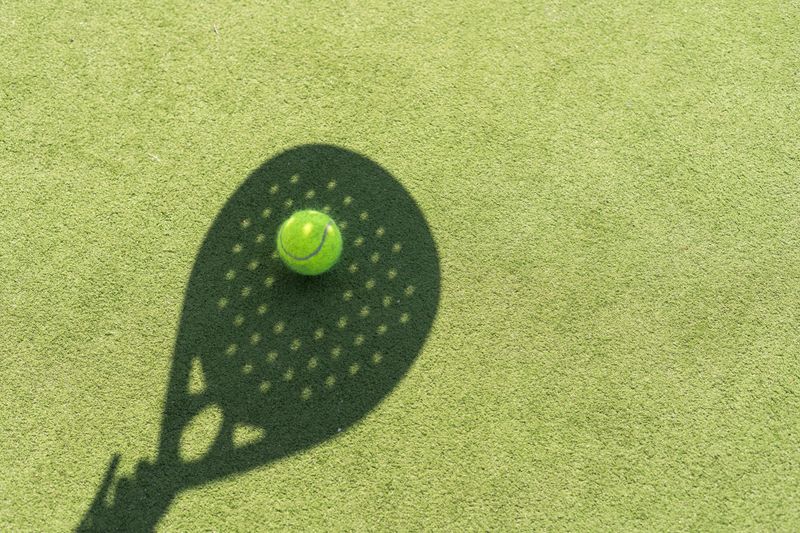 Shadow of a padel racket with a yellow ball on the green grass. High quality photo