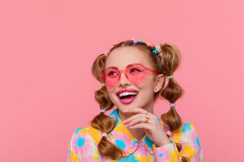 Excited gen Z teen girl with stylish hairstyle with hairpins wearing colorful floral pattern shirt and pink eyeglasses looking away smiling. Studio shot, pink background.