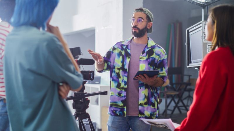 Young Caucasian Male Director With Beard And Glasses, Instructing Diverse Team On Film Set, Holding Tablet, Surrounded By Camera Equipment And Crew In Creative Studio Environment.