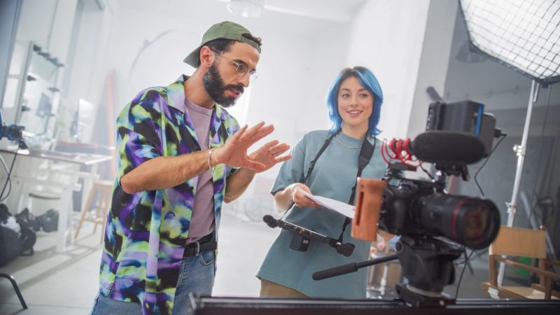 Young Latin Male Director With Beard And Caucasian Female With Blue Hair Discuss Over A Script In A Well-equipped Film Studio, Surrounded By Professional Camera And Lighting Equipment.