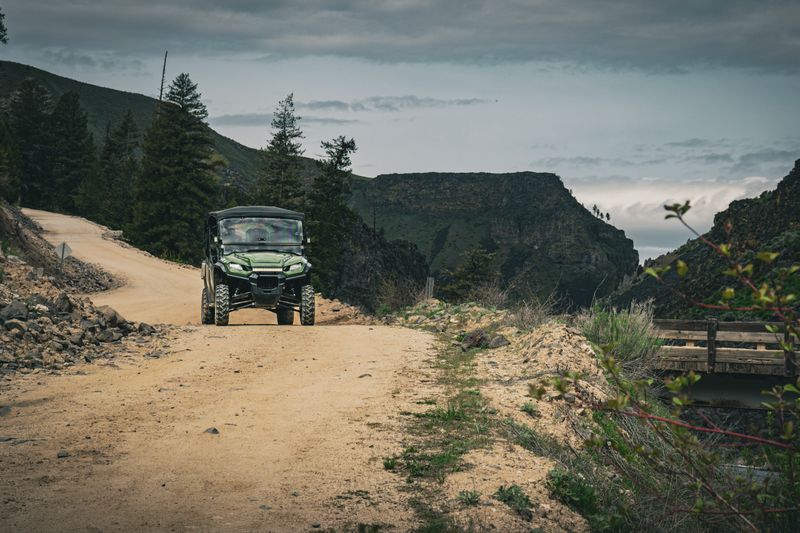 A green UTV is parked on a rugged dirt road set amidst a dramatic mountainous landscape. The overcast sky suggests a moody and adventurous setting.