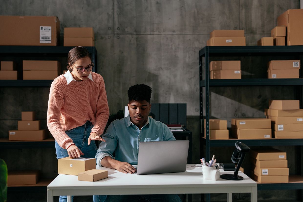 Two people working with packages and a laptop in a storage room.