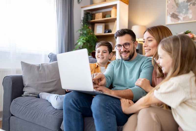 This warm family image showcases a mother, father, and two children engaging with a laptop, promoting a sense of learning and technology at home