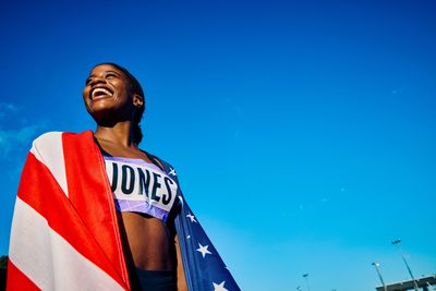 Athlete wrapped in American flag smiling against blue sky.
