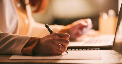 Person writing notes with a pen near a laptop.