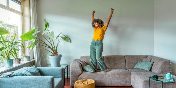 A happy young woman is standing on her couch, arms raised--feeling inspired and powerful.